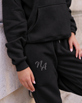 Black Niamh Avenue cuffed joggers featuring a sparkling crystal rhinestone NA logo on the thigh, styled with a matching black hoodie for a luxury casual look.