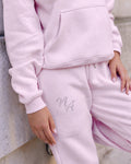 Pink Niamh Avenue cuffed joggers featuring a sparkling crystal rhinestone NA logo on the thigh, styled with a matching black hoodie for a luxury casual look.